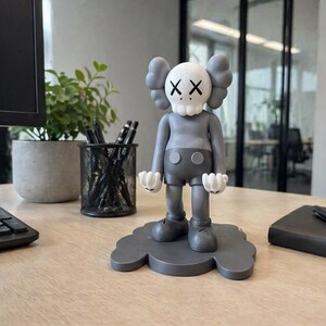 May include: A gray figurine of a cartoon character with a skull-like face and X's for eyes. The figure has gray shorts, shoes, and gloves, and is standing on a gray cloud-shaped base. The figurine is on a wooden desk.