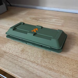 May include: An olive green rectangular case with an orange arrow symbol on the top. The case has a layered design and is resting on a wooden surface. The case appears to be made of plastic.