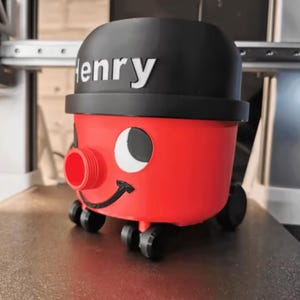 Peut inclure: Un aspirateur fantaisie rouge et noir avec le nom "Henry" sur le dessus. L'aspirateur a un visage souriant, une ouverture circulaire et de petites roues pour la mobilité. Le design rappelle un personnage de dessin animé.