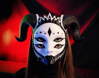 May include: A white and black mask with four eyes and large black horns. The mask has a diamond shape on the forehead and a crown-like design at the top. The mask is worn over the face.