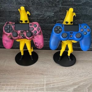 Puede incluir: Dos soportes para mandos de videojuegos con forma de plátanos amarillos, cada uno sosteniendo un mando de diferente color. Un mando es rosa con un patrón de camuflaje, y el otro es azul. Los soportes tienen bases negras.
