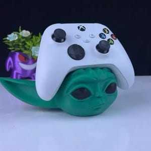 May include: A white Xbox controller sits atop a green Baby Yoda-style figure. The controller features black joysticks and coloured buttons. A purple planter with flowers is in the background.