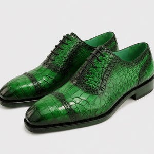 May include: A pair of green leather dress shoes with a textured, crocodile-like pattern. The shoes have black laces and a classic Oxford design. The shoes are displayed on a white background.