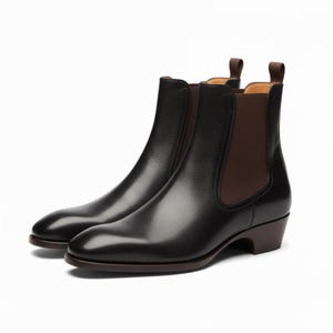 Op de afbeelding: Een paar zwarte leren Chelsea boots met bruine elastische zijpanelen en treklussen. De laarzen hebben een lage hak en een gladde, gepolijste afwerking, perfect voor een klassieke, verfijnde look.