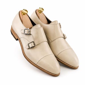 May include: A pair of cream-colored leather double monk strap shoes. The shoes feature two silver buckles, a stitched sole, and a wooden shoe tree inside. The shoes are set against a white background.
