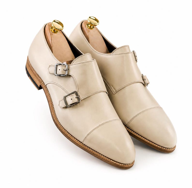 May include: A pair of cream-colored leather double monk strap shoes. The shoes feature two silver buckles, a stitched sole, and a wooden shoe tree inside. The shoes are set against a white background.