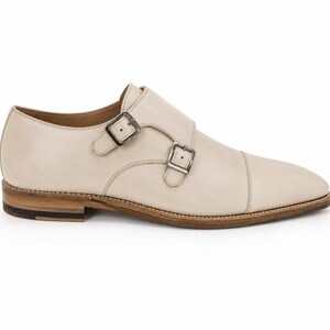 May include: Cream-colored leather double monk strap shoe with two silver buckles. The shoe has a cap toe design and a light brown sole with a dark brown heel. The shoe is a classic style.