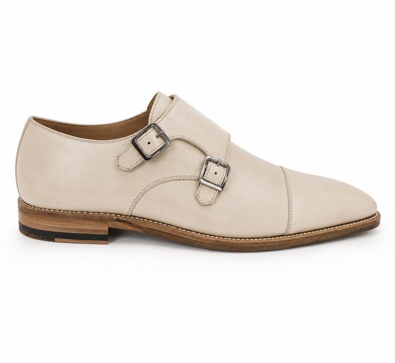 May include: Cream-colored leather double monk strap shoe with two silver buckles. The shoe has a cap toe design and a light brown sole with a dark brown heel. The shoe is a classic style.