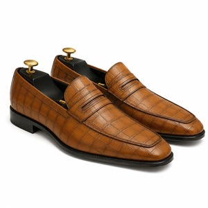 May include: A pair of brown leather penny loafers with a textured, crocodile-like pattern. The shoes have a classic design with a rounded toe and a black sole. Shoe trees are inserted in the shoes.