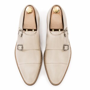 May include: A pair of cream-colored leather dress shoes with double monk straps and silver buckles. The shoes have a pointed toe and are displayed with wooden shoe trees inside. The soles are a light brown color.