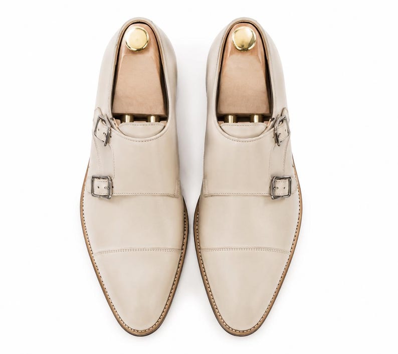 May include: A pair of cream-colored leather dress shoes with double monk straps and silver buckles. The shoes have a pointed toe and are displayed with wooden shoe trees inside. The soles are a light brown color.