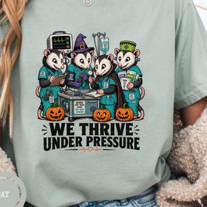 May include: A sage green t-shirt featuring a graphic of four opossums dressed as medical staff in scrubs, with a heart monitor, IV drip, and pumpkins. The text below the graphic reads WE THRIVE UNDER PRESSURE in bold black capital letters.