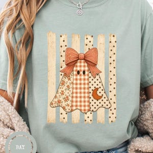 Könnte beinhalten: Ein salbeigrünes Kurzarm-T-Shirt mit einer Grafik eines Geistes, der eine große orangefarbene Schleife trägt. Der Geist ist mit verschiedenen Mustern wie Karos, Punkten und einem Halbmond verziert und vor vertikalen Streifen platziert.