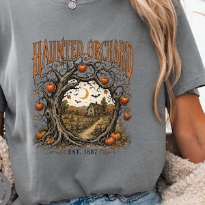 Op de afbeelding: Een grijs t-shirt met korte mouwen en een grafische print van een gedraaide boom die een boog vormt over een landschap met een huis, pompoenen, vleermuizen en een maansikkel. De tekst luidt HAUNTER ORCHARD en EST. 1887 in een oranje lettertype.