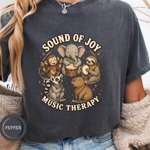 May include: A charcoal gray short-sleeve t-shirt featuring a graphic print of an elephant, monkey, sloth, lemur, and capybara playing musical instruments. The text "SOUND OF JOY" appears above the animals and "MUSIC THERAPY" appears below them.