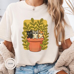 Pode incluir: Uma t-shirt de manga curta na cor marfim com uma estampa gráfica de três gatos sentados dentro de um vaso de flores na cor terracota. Os gatos são preto, cinza tigrado e laranja tigrado. Atrás do vaso há plantas verdes num fundo amarelo.