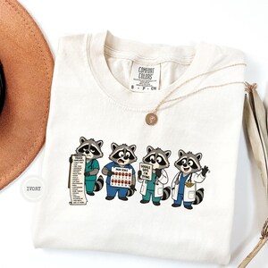 Peut inclure: Un t-shirt ivoire avec un motif de quatre ratons laveurs en dessin animé habillés en professionnels de santé. Les ratons laveurs tiennent une liste de triage, une échelle de douleur et un panneau indiquant Google says I'm dying. Étiquette Comfort Colors.