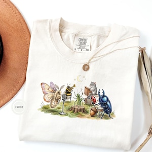 Op de afbeelding: Een ivoorkleurig t-shirt met een afbeelding van een mot, een bij die trompet speelt, een sprinkhaan, een muis achter een piano, een lieveheersbeestje en een kever die drumt. Het shirt heeft een Comfort Colors label in de nek.