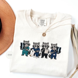 May include: An ivory t-shirt featuring a graphic of four cartoon raccoons dressed as medical professionals. One raccoon holds an X-ray, another wraps a bandage, a third examines a fracture chart, and the fourth points to an X-ray with a pen.
