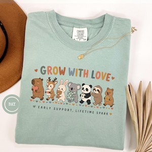 May include: A sage green t-shirt featuring a graphic of various animals including a capybara, deer, rabbit, koala, panda, sloth, and quokka. The text reads "GROW WITH LOVE" and "EARLY SUPPORT, LIFETIME SPARK" with small heart icons in a row.