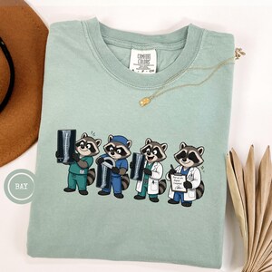 May include: A sage green t-shirt featuring a graphic of four cartoon raccoons dressed as medical professionals. The raccoons are shown holding medical equipment, including X-rays and a clipboard with the text 'Check-up note: Report Completed'.