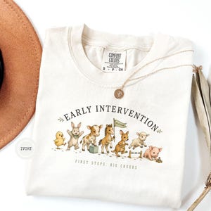 May include: An ivory t-shirt featuring a graphic of various baby animals including a chick, rabbit, calf, pony, kangaroo, lamb, and piglet. The text reads EARLY INTERVENTION and FIRST STEPS, BIG CHEERS. The shirt has a Comfort Colors brand label.