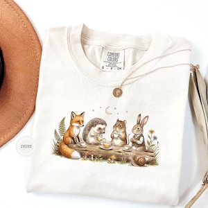 Peut inclure: Un t-shirt ivoire avec une impression graphique représentant un renard, un hérisson, un hamster et un lapin prenant le thé sur une bûche. L'illustration comprend des petits champignons, des fougères, des étoiles et un croissant de lune.