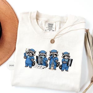 Könnte beinhalten: Ein elfenbeinfarbenes T-Shirt mit einer Grafik von vier Zeichentrick-Waschbären in blauer medizinischer Arbeitskleidung. Die Waschbären sind als orthopädische Chirurgen dargestellt, die chirurgische Instrumente halten, Röntgenbilder untersuchen und einen Eingriff vornehmen.
