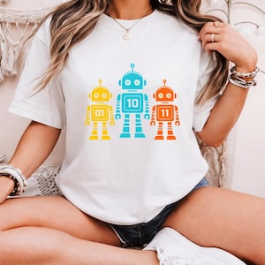 Może przedstawiać: Biała koszulka z trzema kolorowymi wzorami robotów. Roboty są żółte, niebieskie i pomarańczowe, każdy z innym numerem: 01, 10 i 11. Roboty mają anteny i kwadratowe korpusy.