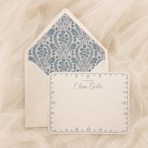 Peut inclure: Une carte de correspondance crème avec une bordure décorative bleue et le texte A NOTE FROM Olivia Carter, accompagnée d'une enveloppe avec un motif floral damassé bleu et blanc sur la doublure intérieure, présentée sur un tissu doux.