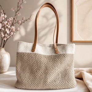 May include: A beige and white woven tote bag with brown leather handles. The bag has a rectangular shape and is made of a textured material. The handles are attached with brass-colored rivets.