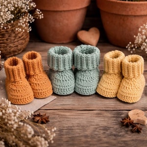 May include: Six pairs of handmade baby booties in shades of orange, seafoam green, and yellow are arranged on a wooden surface. The booties are crocheted with a ribbed cuff design. The background includes terracotta pots and decorative elements.