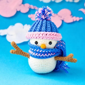 May include: A crocheted snowman with a blue hat and scarf, pink accents, and twig arms. The snowman has a white body, black eyes, and an orange beak. The background is blue with pink and white cloud cutouts.