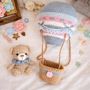 Può includere: Una mongolfiera all'uncinetto con un design a righe blu chiaro e rosa, collegata a un cestino intrecciato. Un piccolo orsacchiotto all'uncinetto con un colletto blu è vicino. Fiori e bottoni decorativi all'uncinetto sono sparsi.