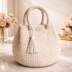 Puede incluir: Bolso de mano de ganchillo color crema con forma redondeada y asas trenzadas. Una borla a juego cuelga de la parte delantera. El bolso tiene una pequeña etiqueta rectangular en la parte superior. El bolso está sobre una superficie de mármol.