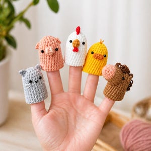 May include: A hand with five crocheted finger puppets: a grey cat, a pink pig, a white chicken, a yellow chick, and a brown horse. Each puppet is designed to fit over a finger, with detailed features, showcasing a farm animal theme.