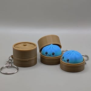 Könnte beinhalten: Drei Schlüsselanhänger mit braunen, zylindrischen Behältern mit Deckel, die an Bambusdämpfer erinnern. Zwei der Behälter sind geöffnet und zeigen eine leuchtend blaue, abgerundete Knödel-Figur mit lächelndem Gesicht, schwarzen Augen und rosa Wangen.
