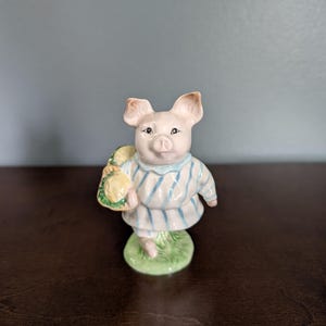 May include: A ceramic figurine of a pig wearing a blue and white striped dress and holding a yellow bag. The pig stands on a green base, with a light blue background. The figurine is a charming decorative item.