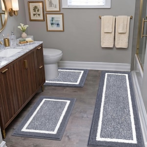 Puede incluir: Un juego de baño con tres alfombras grises con borde blanco. Las alfombras, de forma rectangular y de diferentes tamaños, están diseñadas para colocarse en un baño. El baño cuenta con un tocador de madera y obras de arte enmarcadas.