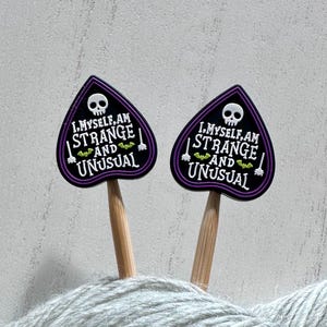 May include: Two black and purple decorative pins with the text "I myself am strange and unusual" and a skull graphic. The pins are on wooden sticks and are placed in a ball of gray yarn.