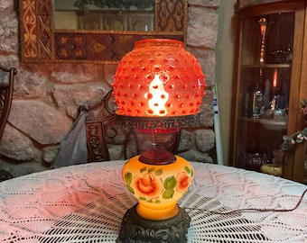 May include: A table lamp featuring a bright red hobnail glass shade and a frosted yellow glass base decorated with hand-painted orange roses and green leaves. The lamp has an ornate dark metal base and a matching metal collar holding the shade.