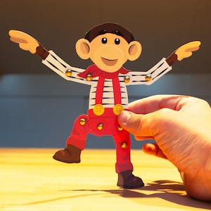 May include: A wooden monkey toy with articulated limbs, wearing a red outfit with suspenders and brown boots. The monkey has a smiling face, brown hair, and large ears. The arms are outstretched, and the toy is held by a hand.