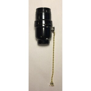 Peut inclure: Une douille de lampe cylindrique noire dotée d'une partie supérieure filetée et d'un mécanisme à chaînette latérale. La chaînette est composée de petites perles métalliques dorées et se termine par un petit embout en forme de cloche dorée.