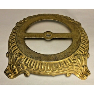 May include: Ornate, antique-style brass-colored metal lamp part. The circular piece features a central opening with a crossbar and decorative floral and scrollwork details. The finish has a slightly aged appearance.