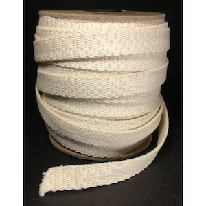 May include: A spool of off-white cotton webbing tape featuring a dense, woven herringbone texture. The fabric strip is tightly wound around a circular cardboard core, with one end of the flat, durable material extending outward from the base.
