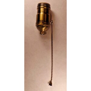 Peut inclure: Un support de lampe en laiton avec une chaîne à tirer. Le corps cylindrique a un dessus fileté et une chaîne à tirer décorative en forme de cloche. Le métal a un aspect légèrement vieilli.
