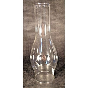 May include: A clear glass lamp chimney with a flared base and a cylindrical top. The chimney has a rounded midsection and is designed to fit over a lamp's flame. The glass is transparent, allowing light to pass through.