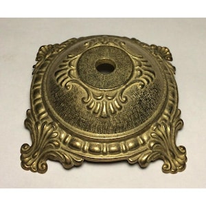 May include: Ornate, antique-style gold-colored metal base with a central hole. The base features detailed scrollwork and a textured finish, suitable for a lamp or decorative item. The base is square with rounded corners.