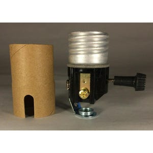 Könnte beinhalten: Eine schwarze und silberne Lampenfassung mit einem schwarzen Knopf und einem Metallsockel. Neben der Fassung befindet sich eine braune Pappröhre. Die Lampenfassung ist für eine Glühbirne konzipiert. Der Hintergrund ist einfarbig grau.