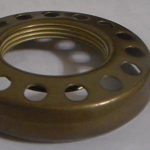 May include: A brass-colored, circular lamp shade holder with a threaded interior and a series of evenly spaced holes around the perimeter. Two small, protruding knobs are visible on the side. This vintage-style component is designed for lighting fixtures.